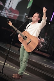A man is standing on a stage, playing a wooden acoustic guitar. He is wearing a white shirt and green pants, smiling and holding a microphone near his mouth. There is a large screen behind him displaying images and text, and the environment suggests a musical or concert setting.