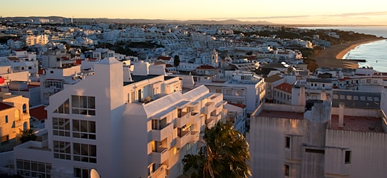 A picturesque coastal cityscape at sunset, with white and light-colored buildings densely packed on sloping hills. The ocean is visible on the right side, with a sandy beach extending along the shoreline. The warm hues of the setting sun cast soft shadows on the buildings, highlighting their geometric shapes and creating a tranquil atmosphere.