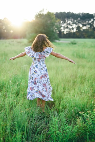 Sunlit outdoor scene with a woman in a flowing dress walking through a field of wildflowers.