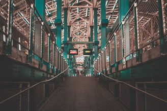 A symmetrical pedestrian walkway with railings on both sides, leading to an elevated structure with signs and intricate metal latticework above. The atmosphere feels urban with industrial elements and teal-colored beams.