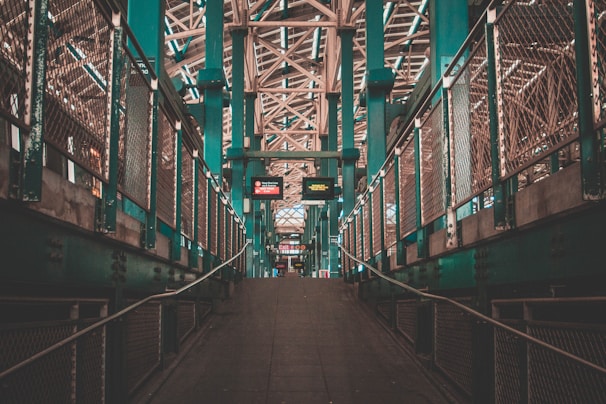 A symmetrical pedestrian walkway with railings on both sides, leading to an elevated structure with signs and intricate metal latticework above. The atmosphere feels urban with industrial elements and teal-colored beams.