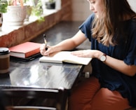 woman sitting in front of black table writing on white book near window