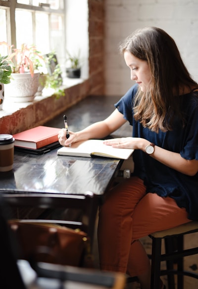 woman sitting in front of black table writing on white book near window