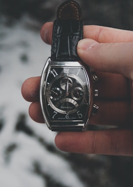 A hand is holding an elegant wristwatch with a dark leather strap. The watch has a silver case with a complex, detailed dial featuring multiple subdials and intricate mechanical components. The background is blurred and consists of soft, muted tones.