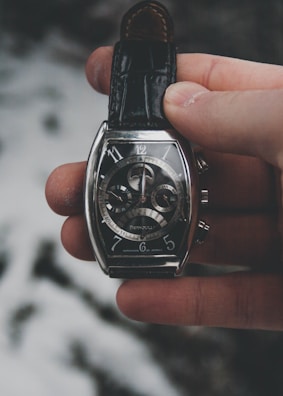 A hand is holding an elegant wristwatch with a dark leather strap. The watch has a silver case with a complex, detailed dial featuring multiple subdials and intricate mechanical components. The background is blurred and consists of soft, muted tones.