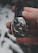 A hand is holding an elegant wristwatch with a dark leather strap. The watch has a silver case with a complex, detailed dial featuring multiple subdials and intricate mechanical components. The background is blurred and consists of soft, muted tones.