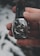 A hand is holding an elegant wristwatch with a dark leather strap. The watch has a silver case with a complex, detailed dial featuring multiple subdials and intricate mechanical components. The background is blurred and consists of soft, muted tones.