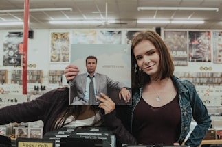 Two people stand in a record store, one holding a record cover in front of their face, aligning it with their body for comedic effect. The other person smiles, standing next to them. Vinyl records are visible in the background, along with store signage and posters on the walls.