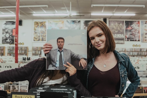 Two people stand in a record store, one holding a record cover in front of their face, aligning it with their body for comedic effect. The other person smiles, standing next to them. Vinyl records are visible in the background, along with store signage and posters on the walls.