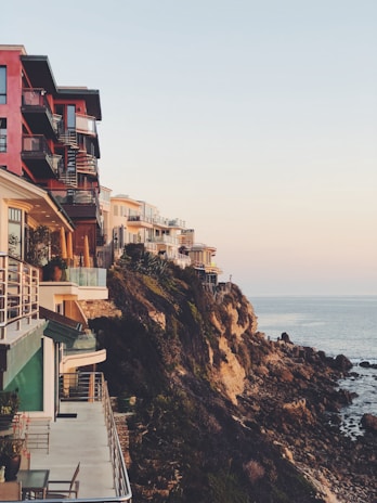 Several modern, multi-story houses are perched on a cliff overlooking the ocean. The architecture features large balconies and terraces, offering expansive sea views. The rocky coastline creates a dramatic contrast with the calm sea and clear sky.
