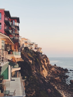 Several modern, multi-story houses are perched on a cliff overlooking the ocean. The architecture features large balconies and terraces, offering expansive sea views. The rocky coastline creates a dramatic contrast with the calm sea and clear sky.