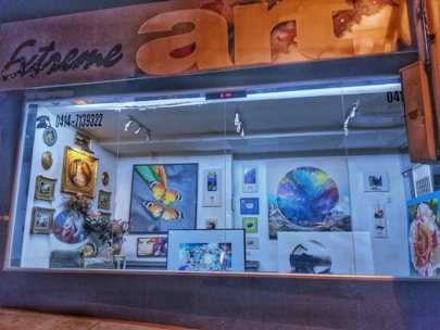 A gallery window display featuring various artworks, including paintings of butterflies, abstract shapes, and natural scenes. The display is well-lit with spotlights and includes floral arrangements and decorative elements. The gallery has a large sign that reads 'art' with the initial letters in a stylized font.