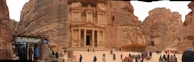 An ancient carved stone facade set within a rocky landscape, bustling with visitors. A small shop on the left displays local wares, while camels are gathered near the structure. The scene presents a blend of historic architecture and present-day activity.