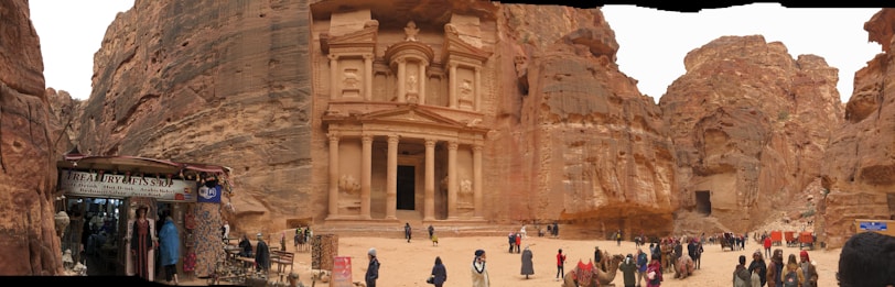 An ancient carved stone facade set within a rocky landscape, bustling with visitors. A small shop on the left displays local wares, while camels are gathered near the structure. The scene presents a blend of historic architecture and present-day activity.