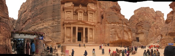An ancient carved stone facade set within a rocky landscape, bustling with visitors. A small shop on the left displays local wares, while camels are gathered near the structure. The scene presents a blend of historic architecture and present-day activity.