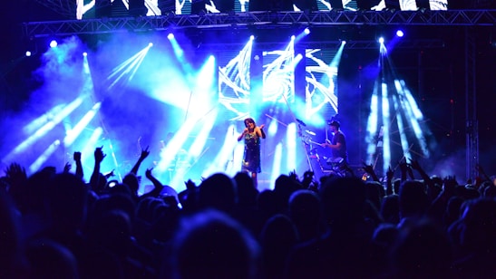 A vibrant concert scene with a crowd facing a brightly lit stage. The stage is illuminated with blue and white lights, and a performer is singing into a microphone while another person plays an instrument. The audience's hands are raised, adding to the dynamic atmosphere.