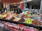 A vibrant market scene featuring a vendor arranging an assortment of colorful candies displayed on a large counter. The varieties include gummies, lollipops, and licorice in various shapes and colors, neatly organized in separate sections. The background shows jars filled with more candy options, enhancing the lively and inviting atmosphere.