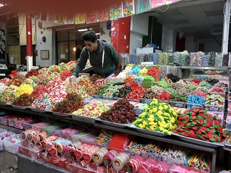 A vibrant market scene featuring a vendor arranging an assortment of colorful candies displayed on a large counter. The varieties include gummies, lollipops, and licorice in various shapes and colors, neatly organized in separate sections. The background shows jars filled with more candy options, enhancing the lively and inviting atmosphere.