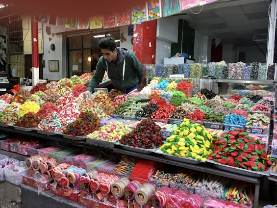 A vibrant market scene featuring a vendor arranging an assortment of colorful candies displayed on a large counter. The varieties include gummies, lollipops, and licorice in various shapes and colors, neatly organized in separate sections. The background shows jars filled with more candy options, enhancing the lively and inviting atmosphere.