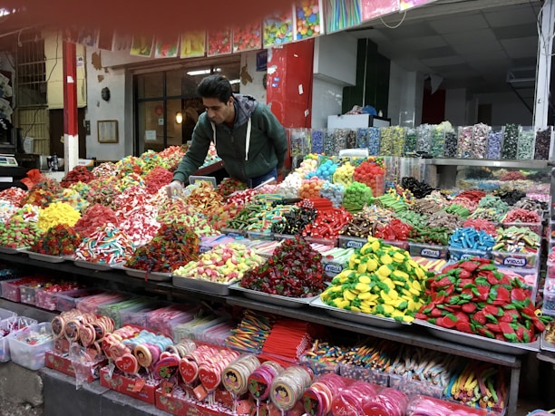 A vibrant market scene featuring a vendor arranging an assortment of colorful candies displayed on a large counter. The varieties include gummies, lollipops, and licorice in various shapes and colors, neatly organized in separate sections. The background shows jars filled with more candy options, enhancing the lively and inviting atmosphere.