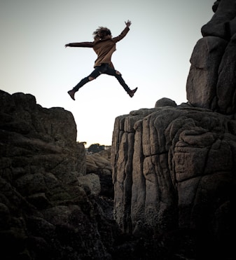 person jumping on big rock under gray and white sky during daytime