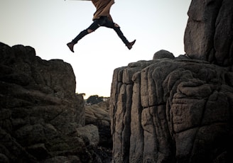 person jumping on big rock under gray and white sky during daytime