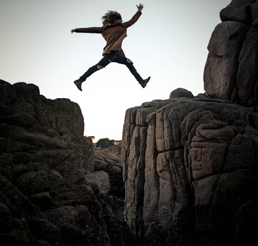 person jumping on big rock under gray and white sky during daytime