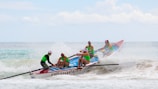 A team of four people in bright green shirts is rowing a colorful boat labeled 'FRESHWATER' through ocean waves under a partly cloudy sky. They are intensely focused, navigating the surf with determination.