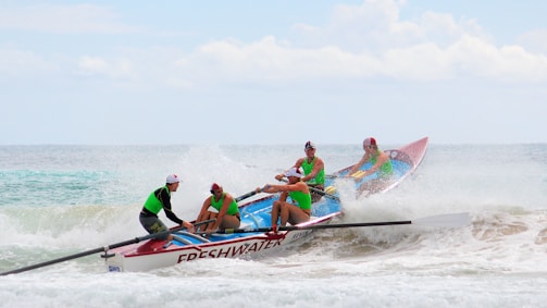 A team of four people in bright green shirts is rowing a colorful boat labeled 'FRESHWATER' through ocean waves under a partly cloudy sky. They are intensely focused, navigating the surf with determination.