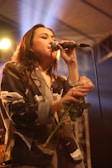 A person is singing into a microphone on stage while holding a rose. The setting appears to be a dimly lit venue with spotlights creating a warm ambiance. The person's attire is a patterned blazer, and there is a sense of concentration and emotion in the performance.