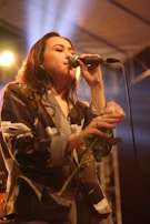 A person is singing into a microphone on stage while holding a rose. The setting appears to be a dimly lit venue with spotlights creating a warm ambiance. The person's attire is a patterned blazer, and there is a sense of concentration and emotion in the performance.
