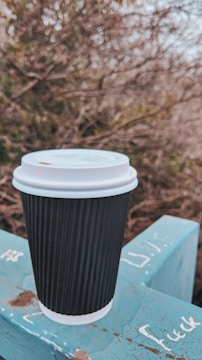 A black ribbed disposable coffee cup with a white lid is placed on a blue surface with some chalk writings. The background consists of out-of-focus foliage and branches, creating a natural setting.
