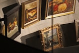 Several framed paintings are displayed in a gallery setting, featuring a variety of artistic styles. The artworks include still life, rural scenes, and landscapes, with each painting surrounded by ornate or simple frames. Shadows are cast on a neutral-colored wall, adding depth to the scene.