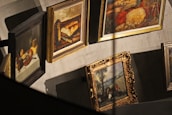 Several framed paintings are displayed in a gallery setting, featuring a variety of artistic styles. The artworks include still life, rural scenes, and landscapes, with each painting surrounded by ornate or simple frames. Shadows are cast on a neutral-colored wall, adding depth to the scene.