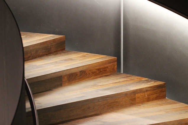 A well-crafted wooden staircase with a smooth, polished finish ascends against a modern, minimalist gray wall. The ambient lighting highlights the texture of the wood and casts subtle shadows.