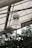A large industrial-style light fixture is hanging from the ceiling of a greenhouse. Metal beams and panels form the structure above, while dense green foliage is visible below, suggesting a lush environment.