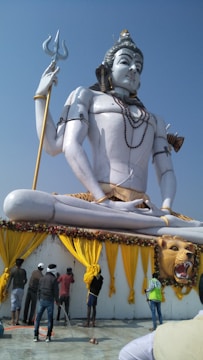 A large statue depicting a deity in a meditative pose with a trident in hand. The statue is white and adorned with jewelry and clothing that includes a leopard print motif. Below the statue, several people are engaged in maintenance activities, possibly cleaning or organizing the area. The surrounding decor includes yellow drapes and floral arrangements, enhancing the grandeur of the scene under a clear blue sky.
