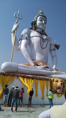 A large statue depicting a deity in a meditative pose with a trident in hand. The statue is white and adorned with jewelry and clothing that includes a leopard print motif. Below the statue, several people are engaged in maintenance activities, possibly cleaning or organizing the area. The surrounding decor includes yellow drapes and floral arrangements, enhancing the grandeur of the scene under a clear blue sky.