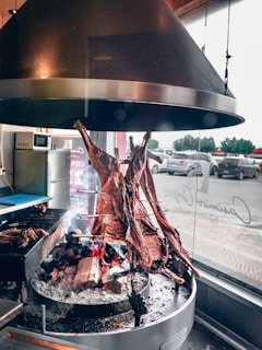 A large vertical grill setup with meat being cooked over an open fire. The grill is located in a restaurant with a window view of parked cars outside. A chimney hood is installed above the grill for ventilation.