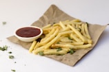 A pile of golden, crispy French fries is placed on a piece of brown paper, garnished with green herbs. A small white dipping bowl filled with red ketchup is positioned beside the fries. The minimalistic background is white, with a few loose herbs scattered around.