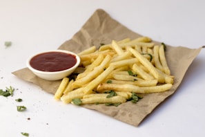 A pile of golden, crispy French fries is placed on a piece of brown paper, garnished with green herbs. A small white dipping bowl filled with red ketchup is positioned beside the fries. The minimalistic background is white, with a few loose herbs scattered around.