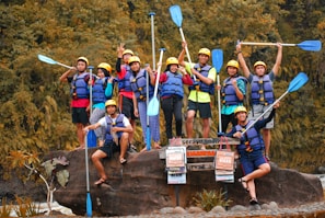 A group of people wearing helmets and life jackets pose cheerfully on a large rock, each holding a paddle. They are surrounded by lush greenery, suggesting a nature or water adventure. Some signs in front of them hint at an outdoor activity location.