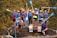 A group of people wearing helmets and life jackets pose cheerfully on a large rock, each holding a paddle. They are surrounded by lush greenery, suggesting a nature or water adventure. Some signs in front of them hint at an outdoor activity location.