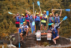 A group of people wearing helmets and life jackets pose cheerfully on a large rock, each holding a paddle. They are surrounded by lush greenery, suggesting a nature or water adventure. Some signs in front of them hint at an outdoor activity location.