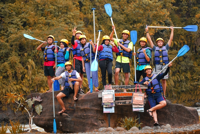 A group of people wearing helmets and life jackets pose cheerfully on a large rock, each holding a paddle. They are surrounded by lush greenery, suggesting a nature or water adventure. Some signs in front of them hint at an outdoor activity location.