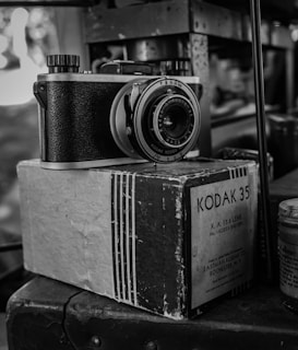 A vintage Kodak 35 camera rests on top of its original box, with intricate details of the camera's body and lens visible. The photograph is in black and white, enhancing the nostalgic feel of the scene.