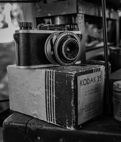 A vintage Kodak 35 camera rests on top of its original box, with intricate details of the camera's body and lens visible. The photograph is in black and white, enhancing the nostalgic feel of the scene.