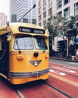 A vintage yellow cable car is traveling along city streets surrounded by tall skyscrapers. American flags are attached to the front of the car, and pedestrians are visible on the sidewalk. The vehicle is marked for the F Market Castro line and bright red tram tracks contrast with the urban landscape.