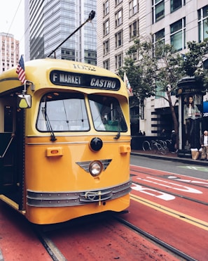 A vintage yellow cable car is traveling along city streets surrounded by tall skyscrapers. American flags are attached to the front of the car, and pedestrians are visible on the sidewalk. The vehicle is marked for the F Market Castro line and bright red tram tracks contrast with the urban landscape.