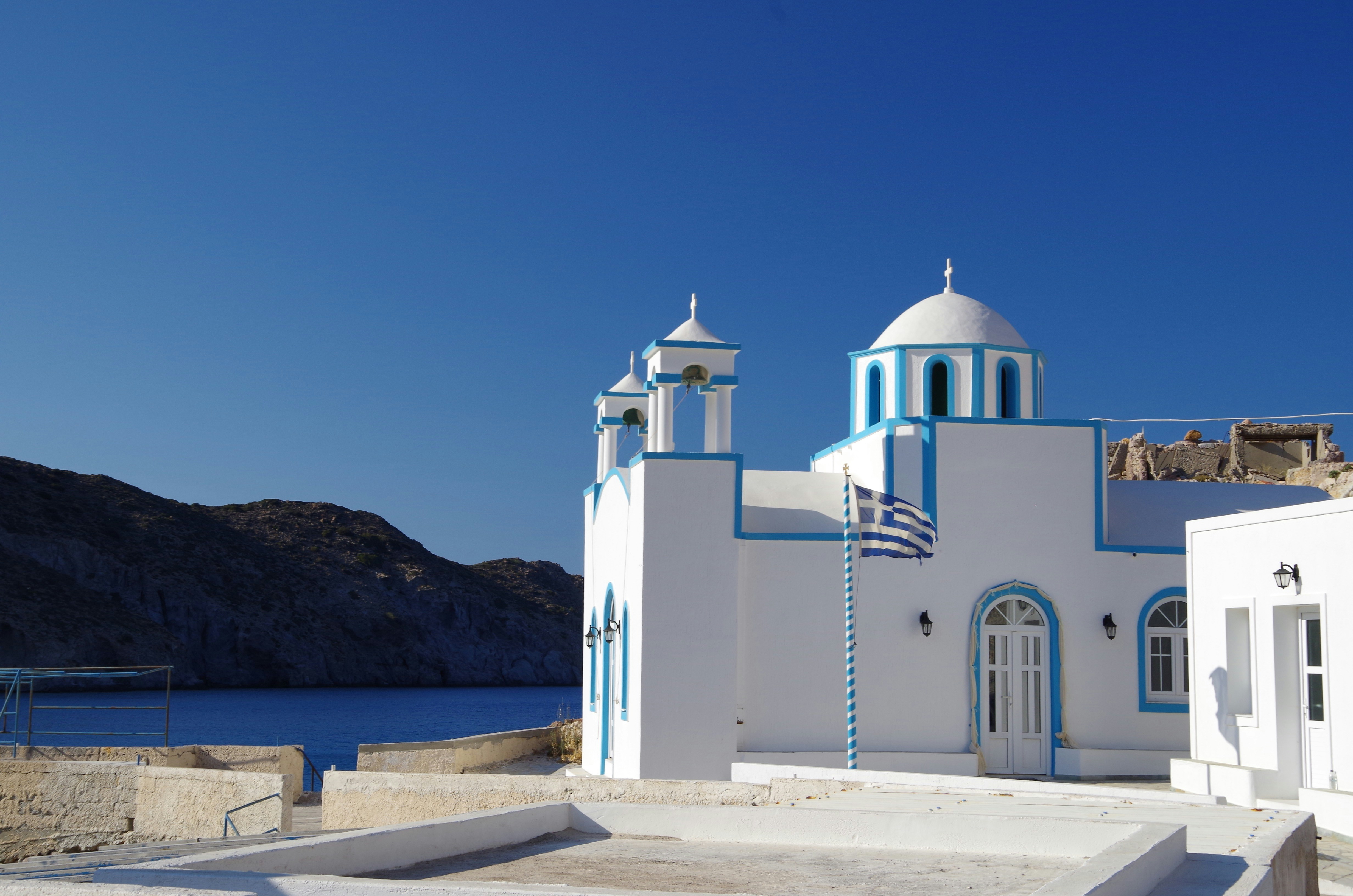 A white building with blue accents and a dome is situated near a coastline, with rocky hills in the background under a clear blue sky. The building features an arched door, several windows, and a striped flagpole with the Greek flag.